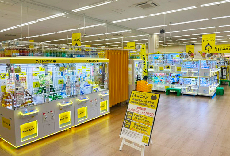 トレルニャン下野店の店内。WonderGOO下野店内に、黄色い照明のクレーンゲーム台が並び、飲料や日用品が景品に入っている。手前に営業時間と未成年の入場時間の案内板